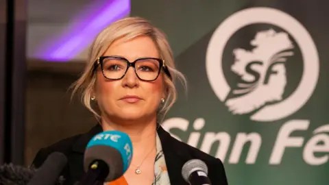 PA Michelle O'Neill مع شعر أشقر تعب ، وارتداء النظارات. إنها تقف أمام الميكروفون.