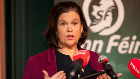 PA Media Mary Lou McDonald ، وهي امرأة ذات شعر بني بطول الكتف ، تتحدث في صف من الميكروفونات الصحفية مع شعار Sinn Fein على الخلفية خلفها