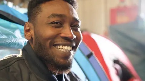 يدعو العمدة إلى كير ستارمر لزيارة معسكرات المهاجرين كاليه 12 Zinki شاب من السودان يعرض بي بي سي خيمته داخل مستودع النبيذ المهجور في كاليه. إنه يبتسم وينظر مباشرة إلى الكاميرا.