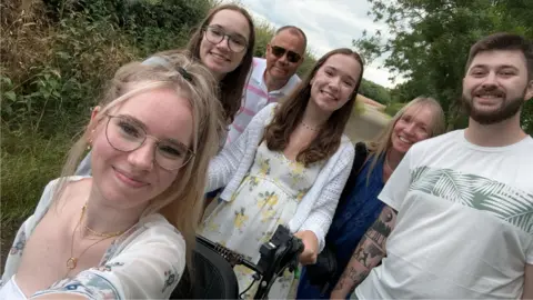 ميغان ديكسون صور عائلية ، بما في ذلك ميغان في المقدمة ، امرأتان ، رجل ، امرأة وأوليفر. إنهم جميعًا ينظرون إلى الكاميرا ويبتسمون ، إحدى النساء يحملن كرسي ميغان المتحرك. هم في الخارج من قبل الحشرات والشجيرات. 