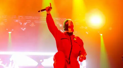 Getty Images DJ Próvaí من Band Kneecap يطرح بذراع واحدة مرفوعة في الهواء ، بينما يرتدي بالاكلافا بألوان العلم الأيرلندي ، على خشبة المسرح في مهرجان كوتشيلا في عام 2025