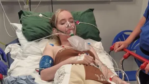 ميغان ديكسون ميغان في سرير المستشفى ، مع الكثير من الأسلاك عليها ، ومع قناع الأكسجين على وجهها. لقد أغلقت عينيها وممرضة بجوارها. 