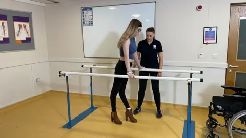 لورا فوستر/بي بي سي ميغان ، التي تمسك ببارين بيضاء ، لمساعدة المشي ، ترتدي سراويل زرقاء وسروال أسود وأحذية بنية. امرأة تقف بجانبها باللون الأسود ، في الغرفة عبارة عن لوحة بيضاء وكرسي متحرك إلى اليمين. 