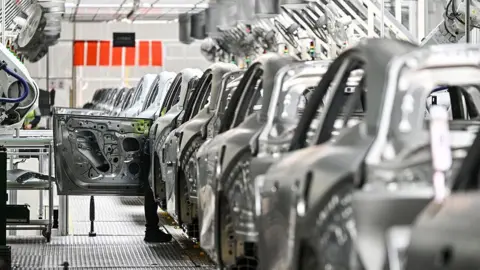 Getty Images يقوم العامل بفحص سيارة كهربائية في مصنع Zeekr في جزيرة ميشان في نينغبو ، في مقاطعة تشجيانغ الشرقية الصينية في 18 أبريل 2025. تظهر الصورة صفًا من المركبات ذات اللون الفضي في مصنع.  