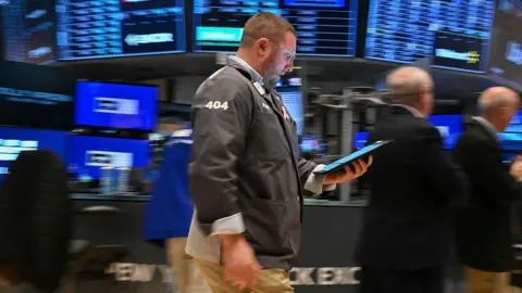 Getty Images يسير المتداول في الماضي وهو يحمل قرصًا على أرضية بورصة نيويورك للأوراق المالية (NYSE) في الجرس الافتتاحي في 21 أبريل 2025 ، في مدينة نيويورك. خلفه شاشات زرقاء غير واضحة توضح الأسواق والرجال في البدلات السوداء. 