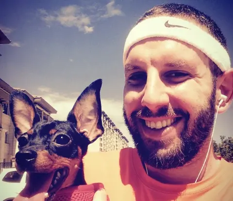 Matthew Radalj Radalj smiling holding a small dog, wearing an orange t-shirt and a white sweatband with earphones in his ears. Behind him the sky is blue and blocks of flats can be seen.