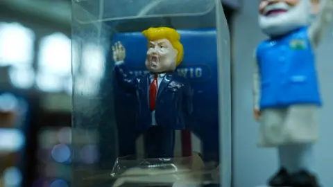 BBC/ XIQING WANG A TOY TRUMP في سترة زرقاء وربطة عنق حمراء في علبة زجاجية في كشك في Yiwu. 