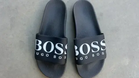 زوج من Hugo Boss Flop Flops التي تم العثور عليها في منزل Pegg. هم أسود ويبدو أنها مصنوعة من الجلد. إنهم مزينون بالكلمات المدرب في رسائل كبيرة ودرب هوغو تحتها. 