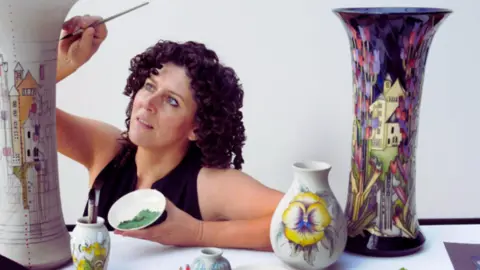 Moorcroft يمكن رؤية امرأة على خلفية بيضاء ، ورسم قطعًا طويلة من الفخار. إنها ترتدي قمة بلا أكمام أسود وهي تحمل وعاء من الطلاء الأخضر.