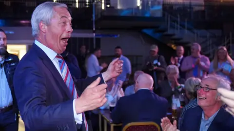 يحتفل PA Media Nigel Farage برفع ذراعيه في الهواء وهو يتطلع إلى حشد في مكان في نيوتن أيكليف ، مقاطعة دورهام. الناس يصفقون ويشاهدونه. إنه يرتدي ربطة عنق مخططة زرقاء وحمراء.