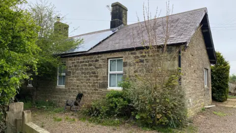 East Cottage عبارة عن كوخ مبني من طابق واحد تقليدي وحوالي 200 عام. هناك شجيرات راسخة بالقرب منها وصفًا مزدوجًا من الألواح الشمسية على السقف المحبب.