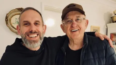 Courtesy Matthew Radalj A smiling, bearded Matthew with his arm around his father, who is also smiling, wearing glasses and a cap. Both are dressed in black. 