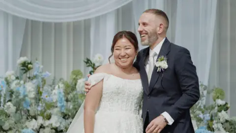 Courtesy Matthew Radalj Radalj on his wedding day in a dark suit with white flowers, with his wife in a white dress. Behind them are white and blue flowers in front of white curtains