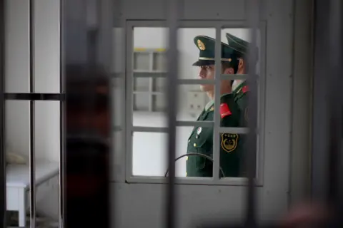 EPA A guard can be seen through bars of a jail door, behind another door. He wears a green uniform with a peaked cap and red on his lapels