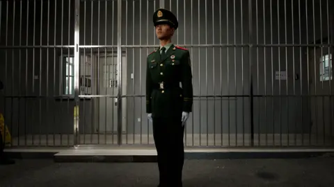 AFP/Getty Images A paramilitary guard stands straight in his green uniform with white gloves in front of a wall of bars. Behind that is a corridor, with a white door and grey walls