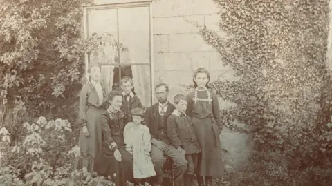 Martin Beal صورة بالأبيض والأسود لمجموعة عائلية تجلس وتقف أمام نافذة. يبدو حتى الآن من أوائل القرن العشرين.