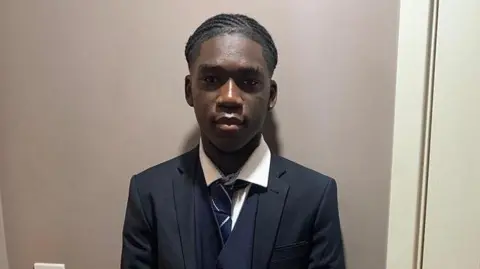 يرتدي ماركوس فاكانا وسائل الإعلام بنسلفانيا بدلة مظلمة وقميصًا أبيض وربطة عنق يقف في الداخل على جدار بيج. إنه يواجه الكاميرا بيديه المغطاة أمامه. تبديل الباب ومفتاح الضوء مرئيًا على كلا الجانبين.