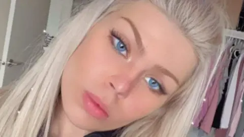 شرطة اسكتلندا شابة ذات شعر أشقر طويل وعيون زرقاء، تنظر إلى الكاميرا، والملابس معلقة خلفها، وباب مفتوح.